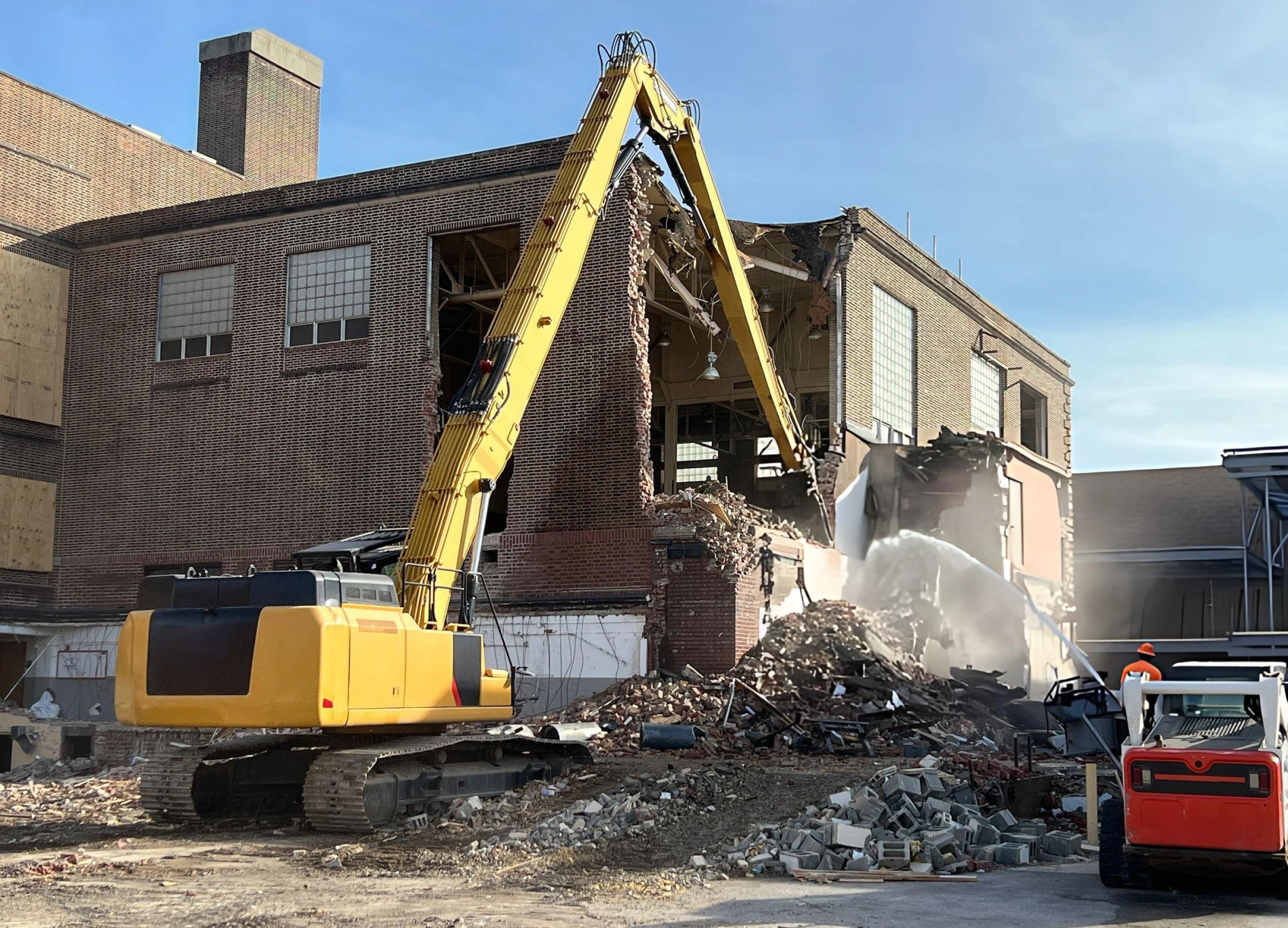 A yellow excavator demolishes a large brick building, with rubble and debris piled in front of the partially collapsed structure under a clear blue sky.