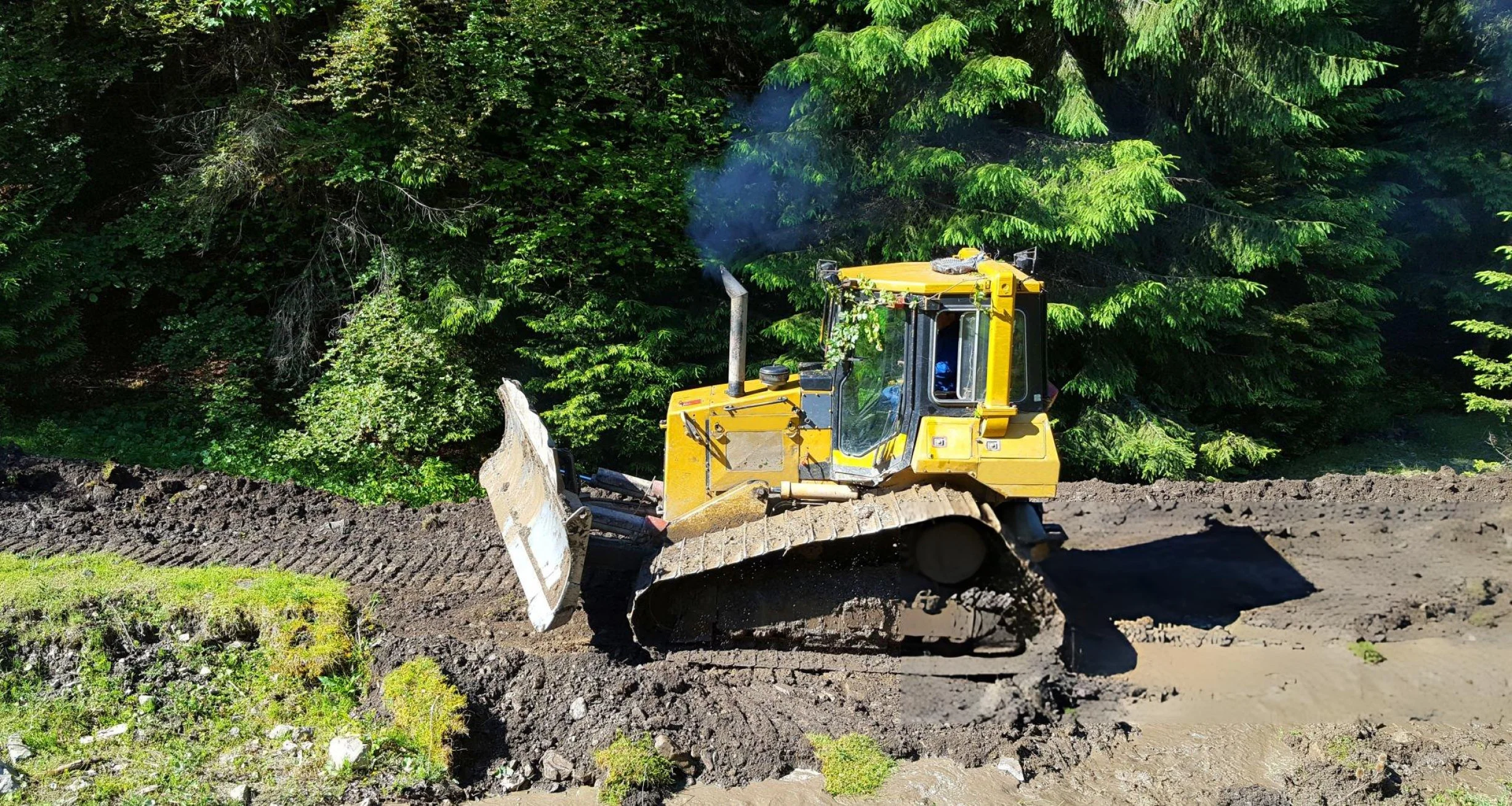 Bulldozer clearing and grading muddy ground in a forested area