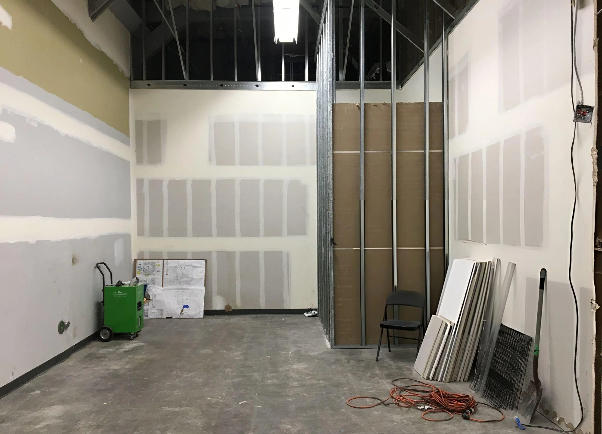 An unfinished commercial interior under construction, with partially finished drywall, exposed metal framing and ceiling structure, a concrete floor, stacked construction materials, a green utility cart, a folding chair, and electrical cords.